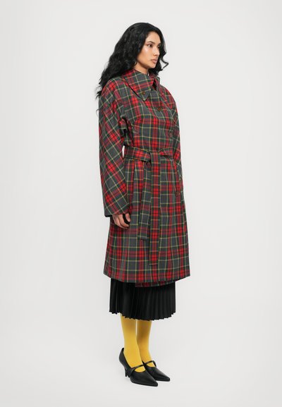 Trench coat a quadri rossi e verdi con cintura, ampio colletto e dettagli con bottoni, abbinato a una gonna nera plissettata e collant gialli.