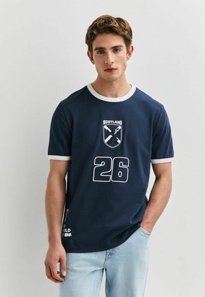 Young man wearing navy blue T-shirt with white trim, Scotland emblem, number 26 print, and light blue jeans, standing against a plain background.