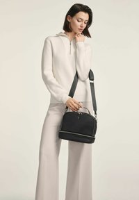 Czarna torba na ramię o eleganckim designie, zapinana na zamek, z podwójnymi opcjami uchwytów. Nosi się na krzyż z białym swetrem w prążki i jasnymi spodniami.