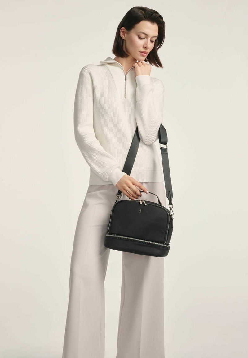 Czarna torba na ramię o eleganckim designie, zapinana na zamek, z podwójnymi opcjami uchwytów. Nosi się na krzyż z białym swetrem w prążki i jasnymi spodniami.