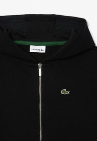 Zwarte zip-up hoodie met een groene binnenvoering, voorzien van een klein wit krokodillenlogo. Gemaakt van zachte stof met geribbelde details.