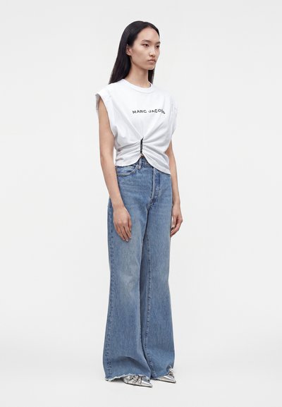 Femme debout en jean bleu à jambes larges, T-shirt sans manches blanc « Marc Jacobs » noué à la taille et chaussures pointues argentées.
