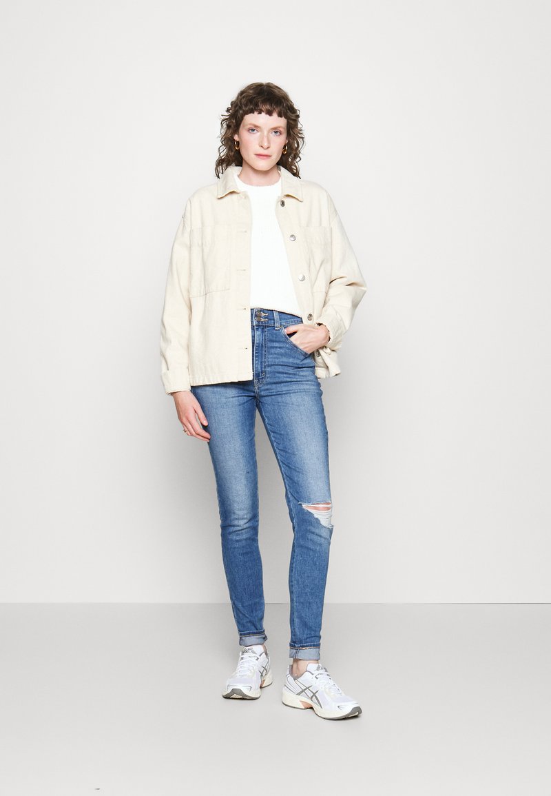 Giacca beige oversize con bottoni sopra un maglione bianco, abbinata a jeans blu attillati con strappi sulle ginocchia e scarpe da ginnastica bianche.