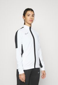 Witte sportjack met rits en zwarte accenten, voorzien van het Nike-logo. Heeft een hoge kraag en een gestroomlijnde pasvorm, gemaakt van rekbaar, lichtgewicht materiaal.