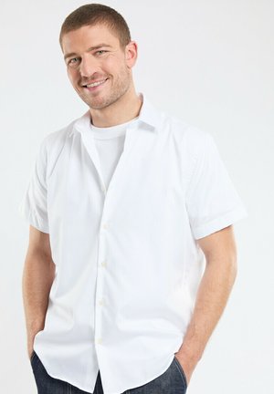 Chemise blanche à manches courtes avec une coupe décontractée, une texture lisse et de petits détails de boutons, portée sur un T-shirt blanc.