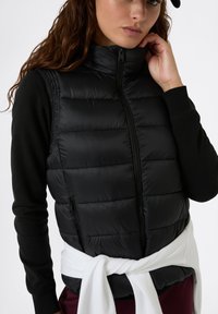 Gilet imbottito nero con colletto alto e zip frontale, caratterizzato da cuciture verticali, indossato sopra un top nero a maniche lunghe, con un dettaglio in cravatta bianca in vita.