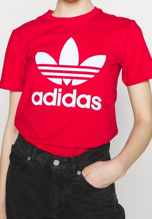 Henkilö, jolla on yllään punainen, lyhythihainen Adidas-paita, jossa on suuri valkoinen trefoil-logo, sekä mustat korkeavyötäröiset farkut, käsi taskussa.