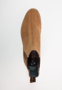 Hnědá semišová kotníková bota s elastickými bočními panely, tvar mandle, kontrastní podšívka a poutko se značkovou etiketou.