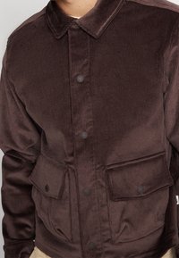 Brun kordflätad jacka med krage, framknappar och två stora fickor. Har en strukturerad textil och en avslappnad passform.