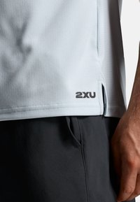 Camiseta deportiva azul claro de manga corta con tela texturizada, aberturas laterales y logo negro "2XU" en la parte inferior derecha. Pantalones cortos negros parcialmente visibles.