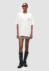 T-shirt oversize color crema con testo nero e motivo a stelle, abbinato a una minigonna texturizzata e stivali neri alti con fibbie decorative.