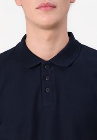 Námořnická modrá polo triko s texturovanou tkaninou, s klasickým límcem a třemi knoflíky na výstřihu. Bez dalších vzorů nebo akcentů.