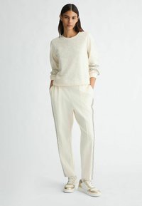 Sweatshirt en tricot crème avec un devant texturé, associé à un jogging crème avec des rayures latérales, et des baskets blanches avec des accents dorés.