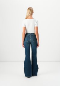 Høyt liv, utsvingte blå denimjeans med en jevn tekstur, to baklommer og en brun label i midjen. Hvit tettsittende topp.