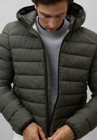 Dunkelgrüne Steppjacke mit Reißverschluss, Kapuze und gestepptem Design. Getragen über einem grauen Pullover, der elastische Bündchen und eine weiche Textur aufweist.