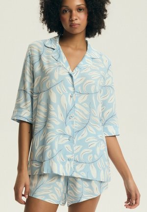 Kobieta w jasnoniebieskim komplecie piżamowym z białym wzorem liści, z krótkimi rękawami i zapięciem na guziki, stojąca na jednolitym tle.