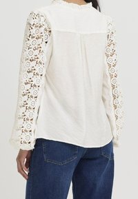Blouse blanche en tissu texturé avec manches longues en dentelle. Le col présente un détail volanté, et le dos est orné de coutures verticales.