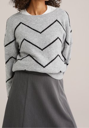 Vrouw die een grijze trui met zwarte zigzagstrepen draagt, ingestopt in een hooggetailleerde donkergrijze rok, tegen een effen achtergrond.