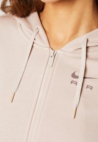 Ljusrosa zip-up hoodie med en texturerad tyg, som har en huva, snörning och en liten mörkgrå logotyp på vänstra sidan.