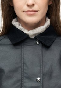 Κοντινό πλάνο ενός ατόμου που φοράει σκουρόχρωμο σακάκι με μαύρο γιακά από κορντουρό πάνω σε λευκό πλεκτό πουλόβερ με ζιβάγκο.