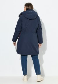 Námořnická modrá parka se staženým pasem, kapucí a výšivkou loga. Disponuje bočními kapsami a hladkým, voděodolným povrchem.