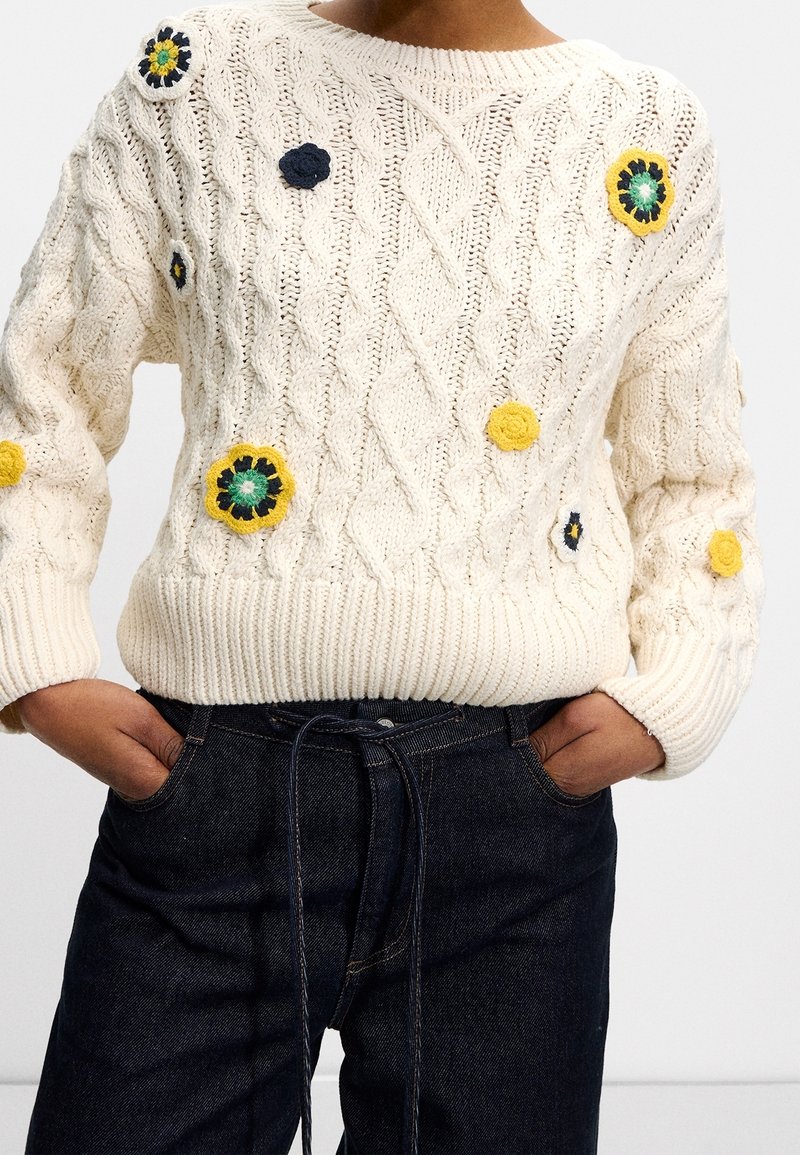 Pull blanc en maille torsadée avec des patchs de fleurs colorées au crochet, porté avec un jean bleu foncé, mains dans les poches.