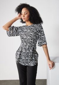 Schwarz-weißer Oberteil mit Zebramuster, lockeren Ärmeln und taillierter Taille, kombiniert mit schwarzen Hosen. Glattes Material, lässige Silhouette.