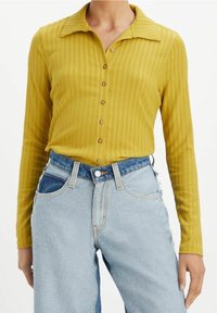 Långärmad skjorta i senapsgul färg med ribbat mönster, kragställd hals och knappar framtill; buren med ljusblå denimjeans med en urtvättad design.