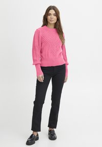 Élénk rózsaszín kötött pulóver texturált geometrikus mintával, bordás nyakkal és mandzsettával, fekete slim-fit nadrággal és fekete loaferekével párosítva.