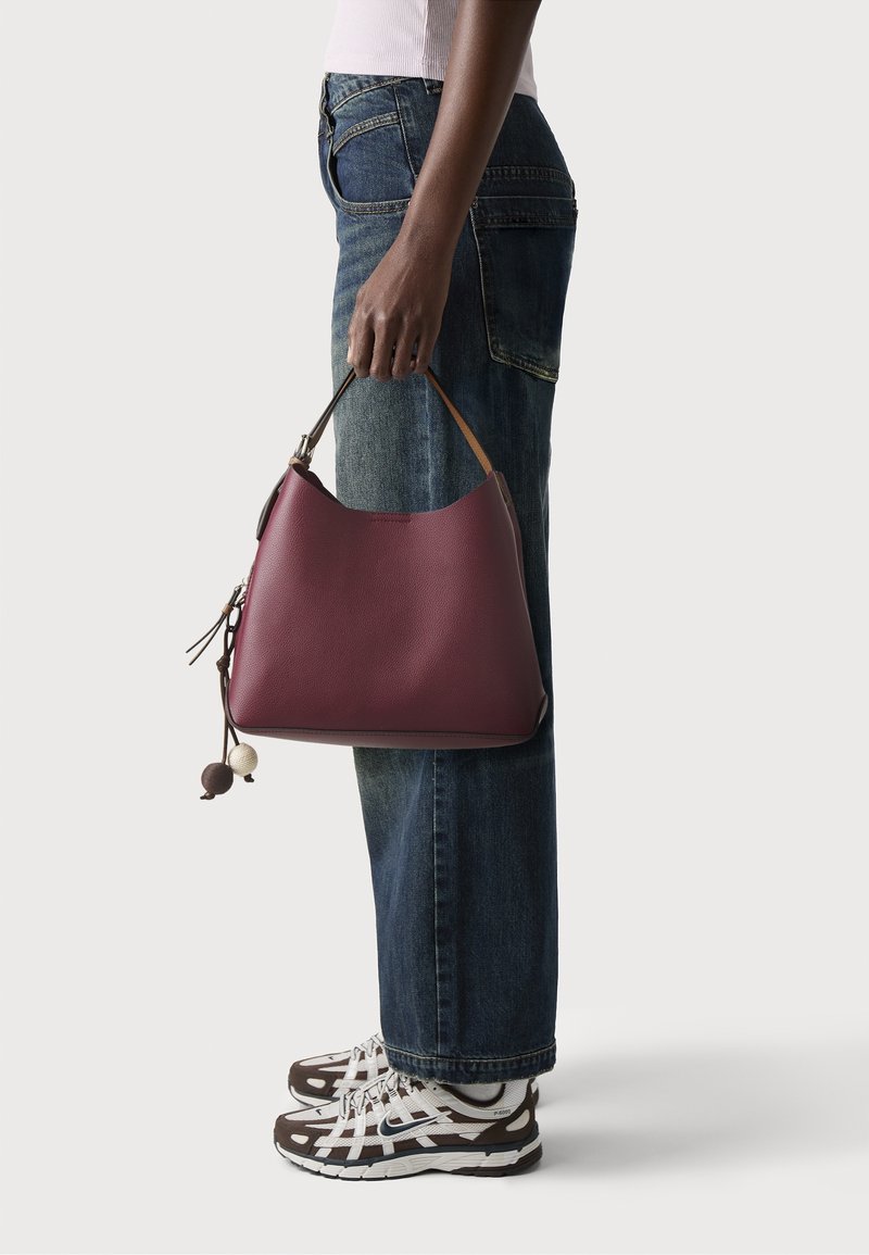 Personne en jean bleu et haut blanc tenant un sac à main en cuir bordeaux avec des pompons décoratifs, portant des baskets blanches et marron.