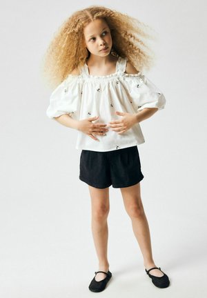 Mladá dívka s kudrnatými vlasy, oblečená v bílém topu s odhalenými rameny, černých šortkách a černých botách, stojící s rukama u pasu.