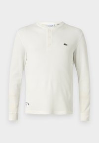 Langärmliges cremefarbenes Rippen-Henley-Shirt aus weicher Baumwolle, mit einer Drei-Knopf-Leiste und einem kleinen grünen gestickten Logo auf der Brust.
