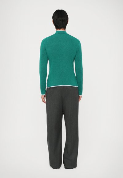 Pullover verde de malha com mangas longas e gola alta, combinado com calças largas plissadas de cor cinza escuro. O pullover tem uma textura suave.