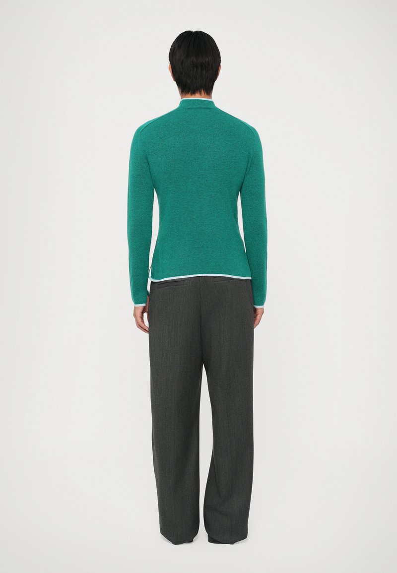 Suéter de punto verde con mangas largas y cuello alto, combinado con pantalones holgados de plisado en gris oscuro. El suéter tiene una textura suave.