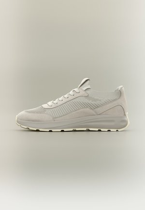 Zapatilla deportiva beige clara con parte superior de punto y ante, cierre de cordones y suela acolchada sobre fondo neutro.