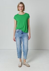 Femme aux cheveux blonds courts, portant un t-shirt vert à manches courtes, un jean bleu clair et des baskets beiges, debout devant un fond uni.