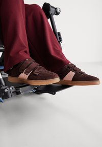 Καφέ σουέτ παπούτσια με βέλκρο λουράκια, διακοσμημένα με ροζ λεπτομέρεια και γκρι σόλα. Τοποθετημένα σε υποπόδιο αμαξιδίου, με μοβ παντελόνια ορατά.