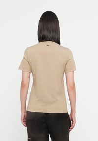 Maglietta beige a maniche corte in cotone; presenta un piccolo logo verde sulla parte posteriore vicino al collo. Tessuto liscio e vestibilità classica.
