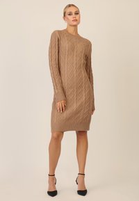 Strickkleid in Hellbraun mit Rautenmuster, Rundhalsausschnitt, langen Ärmeln und geripptem Saum. Getragen mit schwarzen High Heels.