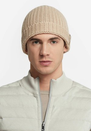 Beżowa dzianinowa czapka o fakturze ściągacza, o zaokrąglonym kształcie i delikatnym metalowym akcentem. Noszona przez osobę w jasnoszarym swetrze z zamkiem.