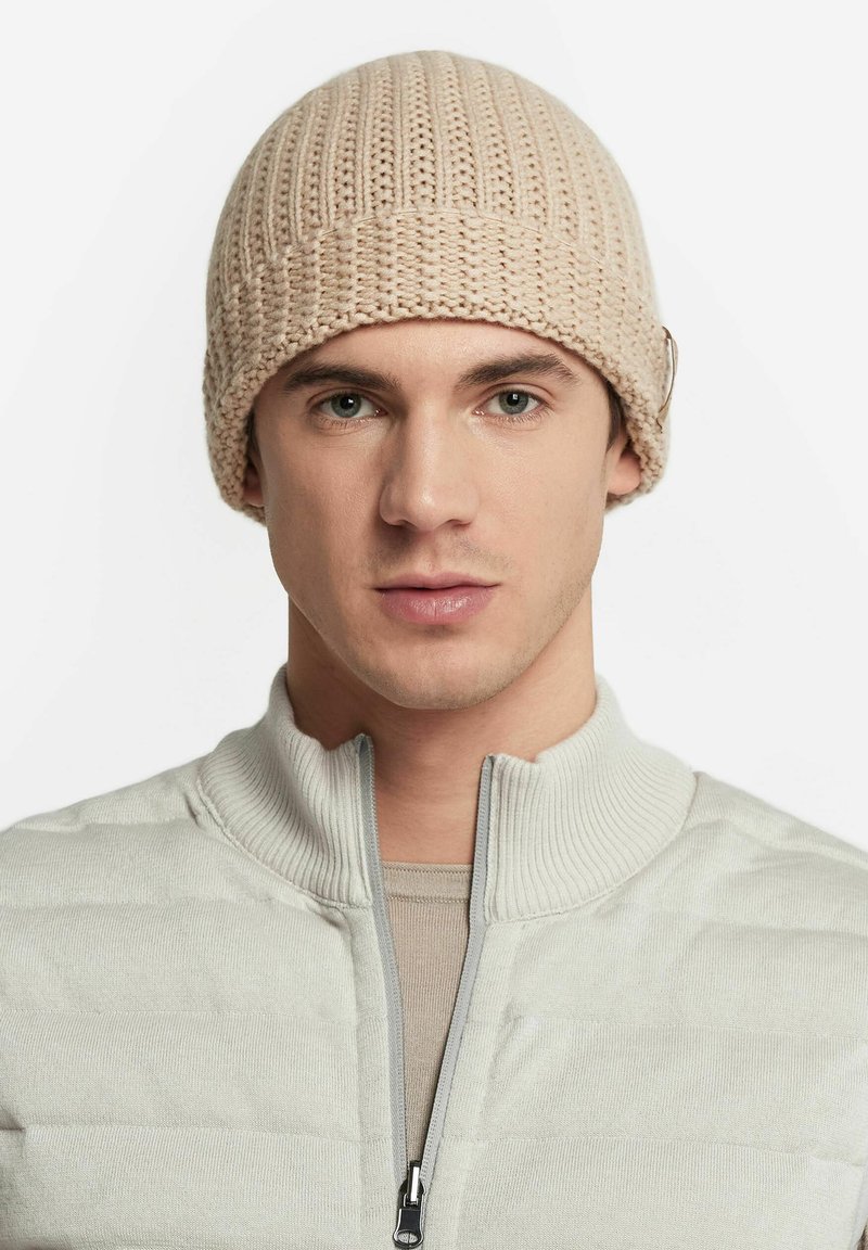 Berretto a maglia beige con texture a costine, forma arrotondata e un sottile accento metallico. Indossato da una persona in un maglione zip grigio chiaro.