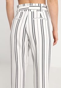 Bílé pruhované kalhoty s černými svislými pruhy, vysokým pasem a dvěma zadními kapsami. Hladká textura a upravený střih.