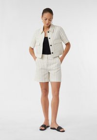 Creme gestreifte kurzärmelige Jacke mit schwarzen Knöpfen und einer Tasche, kombiniert mit passenden Shorts. Getragen über einem schwarzen Oberteil und schwarzen Rutsch-Sandalen.