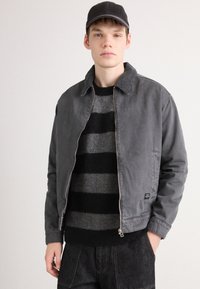 Young man wearing a black cap, grey zip-up jacket, and black-and-grey striped sweater, standing with one hand in his pocket.