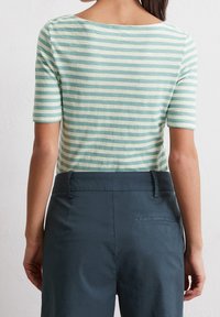 Mujer vestida con una blusa de rayas verdes y blancas de manga corta metida en pantalones de tiro alto de color azul oscuro, de pie contra una pared blanca lisa.