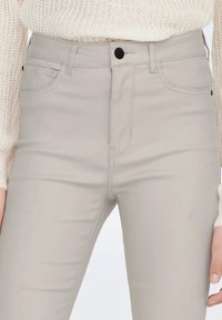 Lichtbeige hoge taille jeans met een gladde textuur. Beschikt over voorzakken, riemlussen en een sluiting met een knoop aan de voorkant. Stiksel details duidelijk zichtbaar.