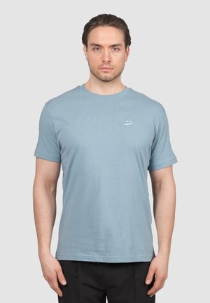 Mężczyzna stojący przodem, ubrany w jasnoniebieską koszulkę z krótkim rękawem, z małym białym logo uśmiechniętej buźki na piersi.