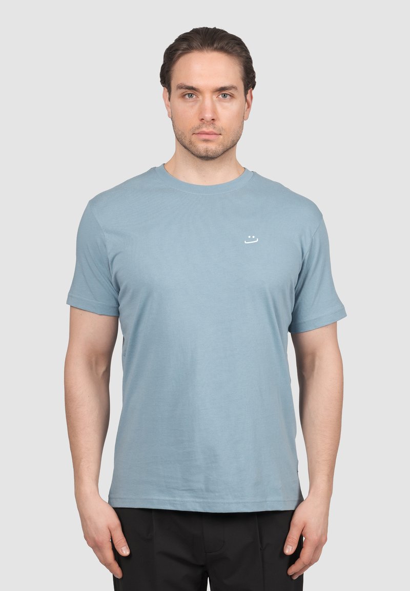 Mees seisab näoga ettepoole, kandes helesinist lühikese varrukaga T-särki, mille rinnal on väike valge naeratava näo logo.