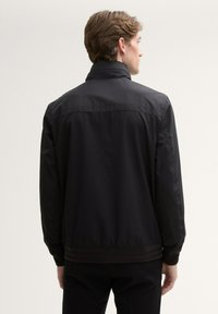 Zwarte waterdichte jas met een kraag, elastische manchetten en tailleband. Gestructureerde stof met scherpe lijnen en minimale naden, weergegeven vanuit de achterkant.
