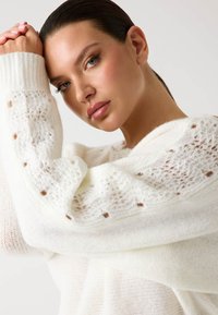 Pull en laine blanche tricoté avec des manches texturées en dentelle et de petits points bruns. Tissu doux, coupe décontractée et large encolure.